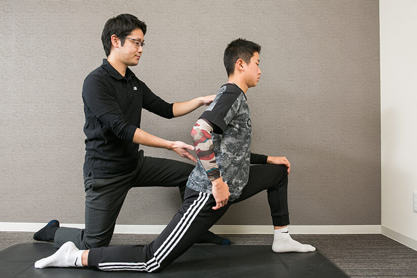 片膝立ちの姿勢を横から撮り、施術者が肩と骨盤に手を添えて指導しているところ