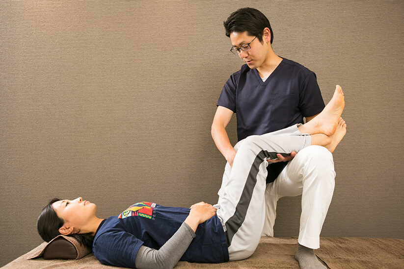 仰向けの姿勢で脚のつけ根に手を当てる施術の場面