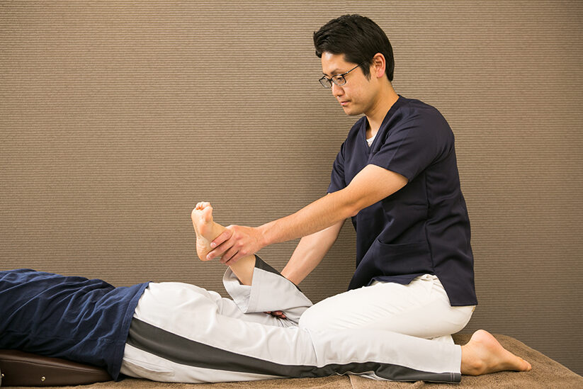 うつ伏せに寝た方の足を持って、足首の動きを確かめているところ
